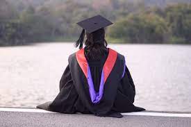 A person dressed in a graduation cap and gown sits with their back facing the camera.
